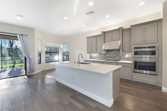 kitchen featuring black electric stovetop, a kitchen island with sink, double oven, sink, and gray cabinets