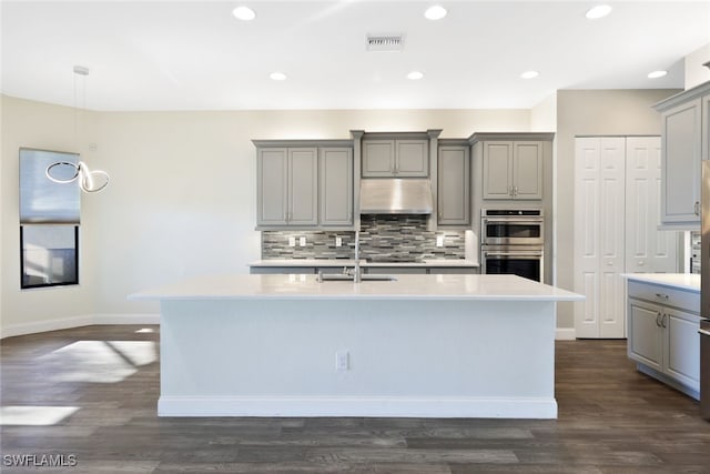 kitchen featuring gray cabinetry, sink, stainless steel double oven, dark hardwood / wood-style flooring, and a center island with sink