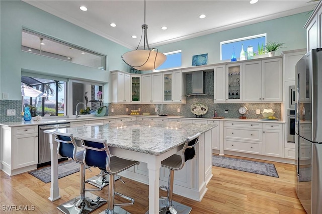 kitchen featuring stainless steel appliances, crown molding, wall chimney range hood, white cabinetry, and hanging light fixtures