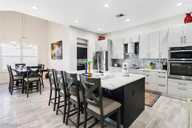 kitchen featuring a kitchen bar, wall chimney range hood, an island with sink, and hanging light fixtures