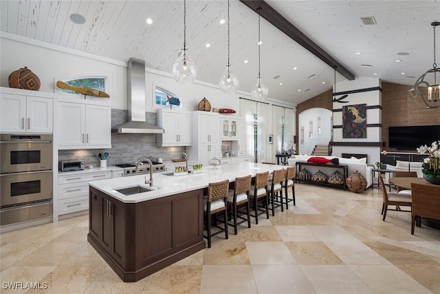 kitchen featuring pendant lighting, wall chimney exhaust hood, a center island with sink, and high vaulted ceiling