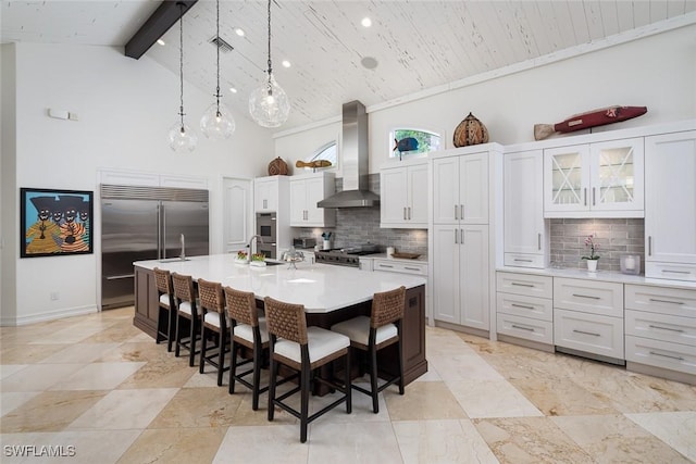 kitchen with appliances with stainless steel finishes, tasteful backsplash, wall chimney exhaust hood, beam ceiling, and an island with sink