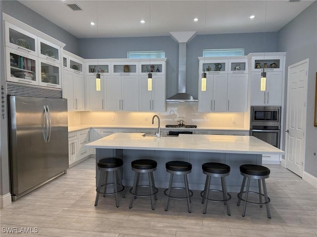 kitchen with white cabinetry, stainless steel appliances, wall chimney range hood, pendant lighting, and a kitchen island with sink