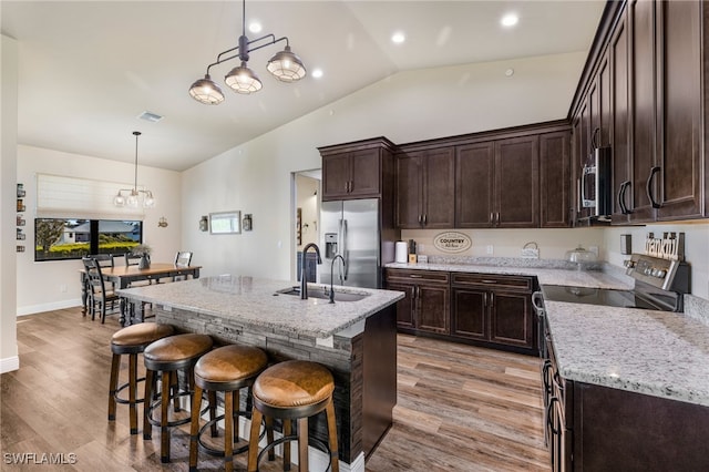 kitchen with a center island with sink, sink, hanging light fixtures, light stone countertops, and stainless steel appliances