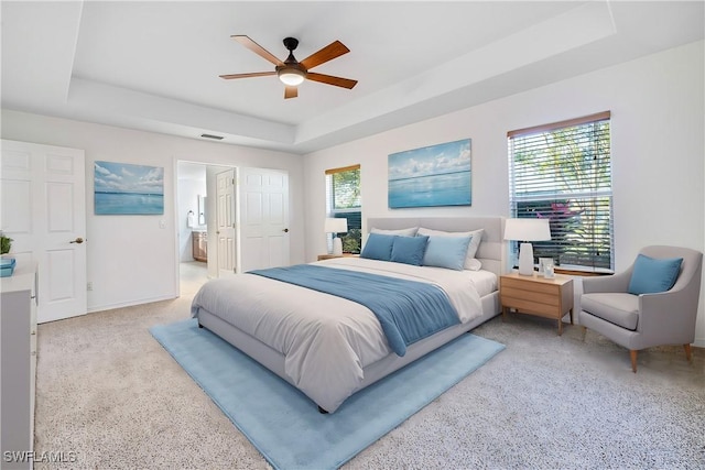 bedroom with ceiling fan, light colored carpet, ensuite bathroom, and a tray ceiling