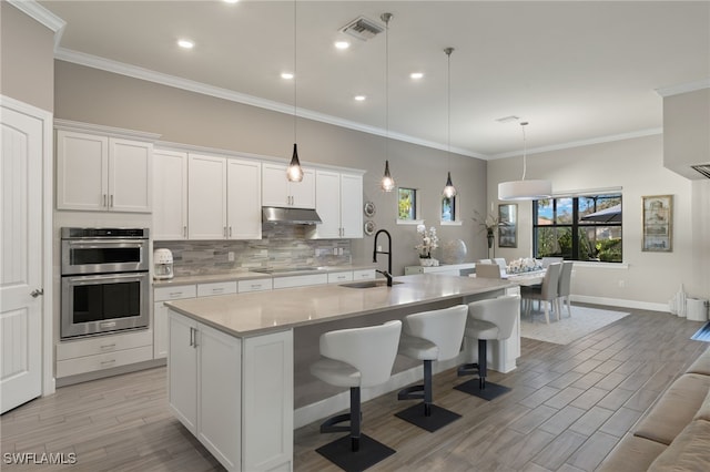 kitchen featuring a center island with sink, black electric stovetop, sink, stainless steel double oven, and white cabinetry