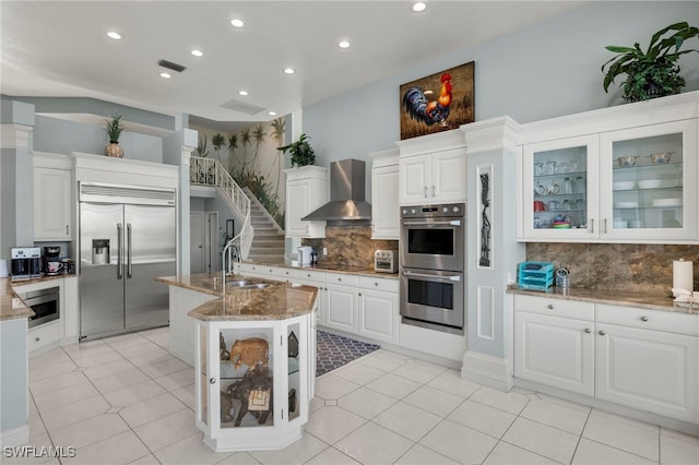 kitchen featuring sink, built in appliances, wall chimney exhaust hood, light stone countertops, and white cabinetry