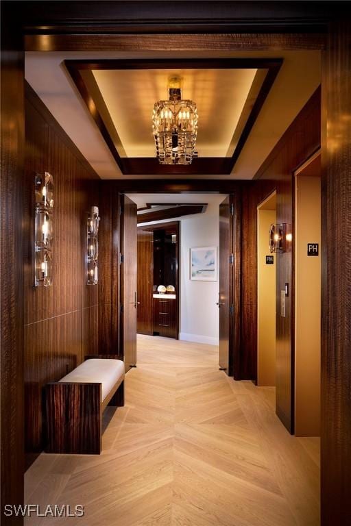 corridor with light parquet flooring, an inviting chandelier, a raised ceiling, and wooden walls