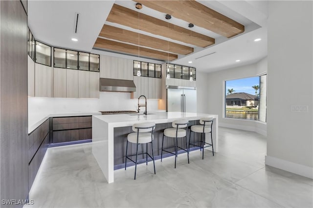 kitchen featuring sink, built in refrigerator, hanging light fixtures, ventilation hood, and an island with sink