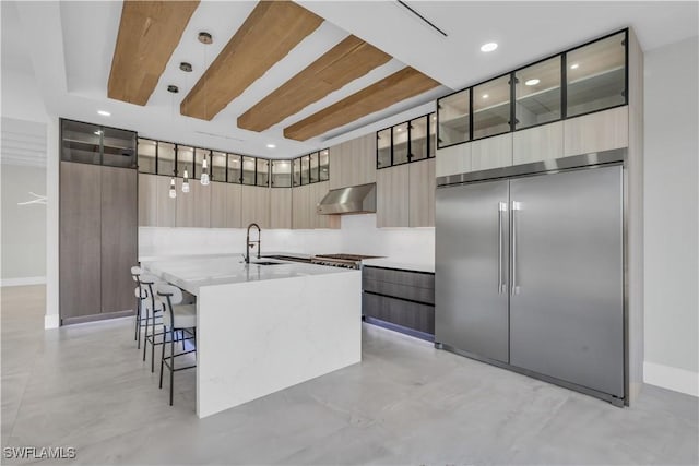 kitchen with built in fridge, light brown cabinetry, decorative light fixtures, sink, and a center island with sink