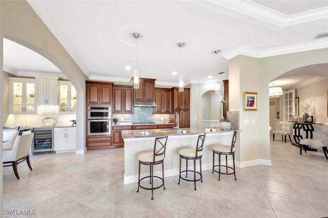 kitchen with stainless steel double oven, under cabinet range hood, beverage cooler, cooktop, and a kitchen bar