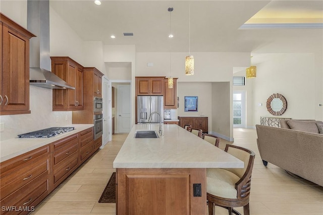 kitchen featuring a kitchen bar, stainless steel appliances, a kitchen island with sink, decorative light fixtures, and wall chimney range hood