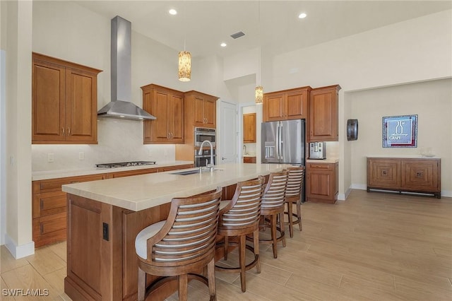 kitchen featuring stainless steel appliances, a kitchen island with sink, hanging light fixtures, wall chimney exhaust hood, and sink