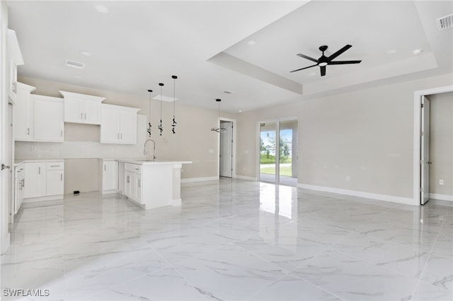 kitchen with a raised ceiling, ceiling fan, white cabinetry, hanging light fixtures, and an island with sink