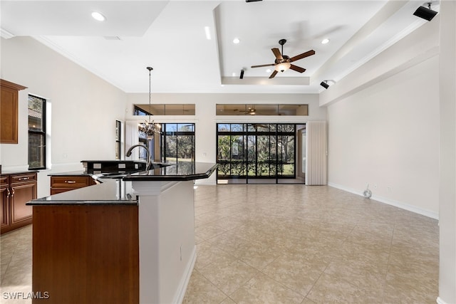 kitchen with pendant lighting, ceiling fan with notable chandelier, sink, an island with sink, and a tray ceiling