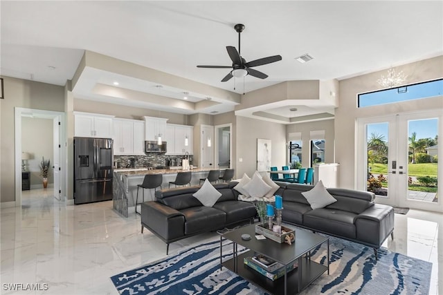 living room with ceiling fan with notable chandelier, a raised ceiling, and french doors