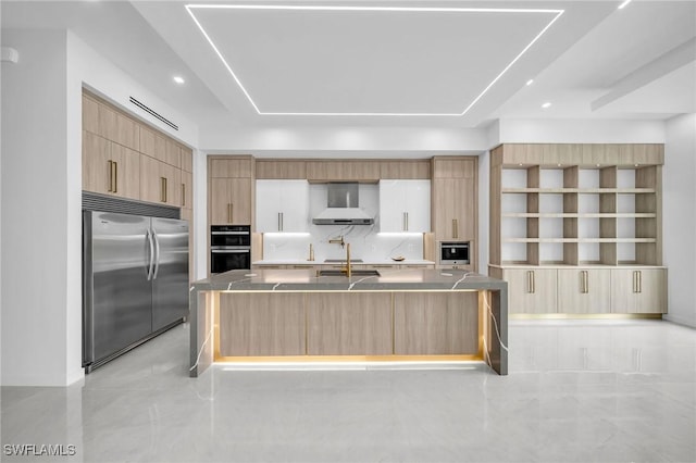 kitchen featuring a kitchen island with sink, wall chimney range hood, double oven, tasteful backsplash, and stainless steel built in refrigerator