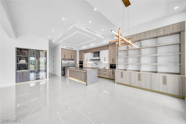 kitchen with stainless steel fridge, wall chimney range hood, light brown cabinets, decorative light fixtures, and a kitchen island