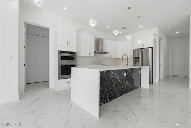 kitchen with appliances with stainless steel finishes, wall chimney exhaust hood, a kitchen island with sink, decorative light fixtures, and white cabinetry