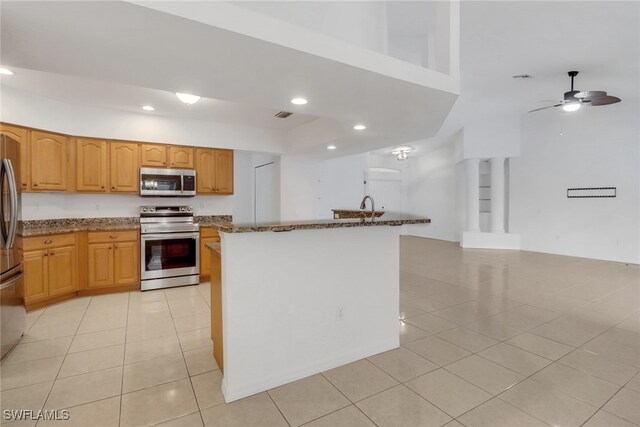 kitchen featuring light tile patterned floors, a center island with sink, ceiling fan, stainless steel appliances, and dark stone counters