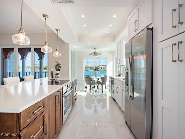 kitchen with a raised ceiling, stainless steel appliances, hanging light fixtures, ceiling fan, and white cabinetry