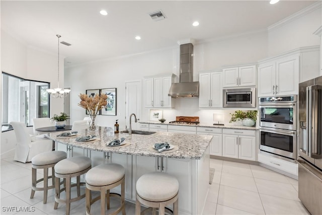 kitchen featuring a kitchen island with sink, white cabinets, wall chimney range hood, hanging light fixtures, and appliances with stainless steel finishes