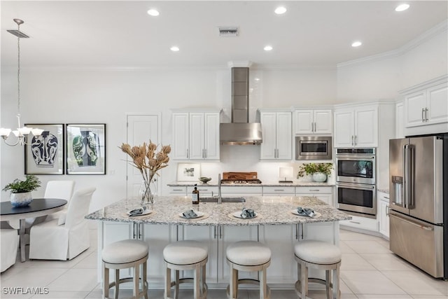 kitchen featuring stainless steel appliances, a kitchen island with sink, wall chimney range hood, white cabinets, and hanging light fixtures