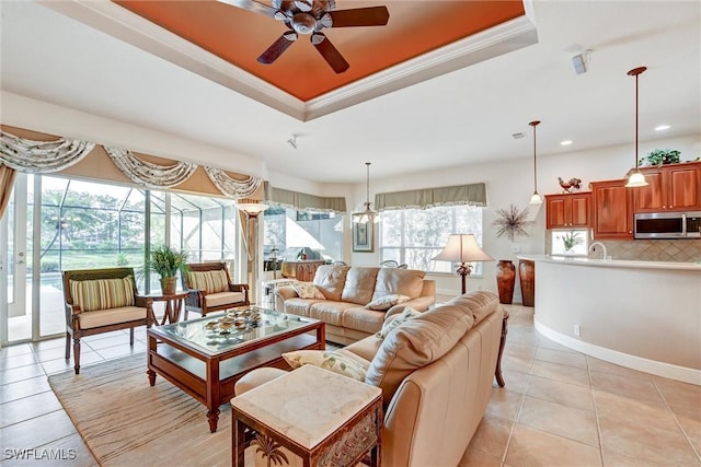 living room with a tray ceiling, crown molding, light tile patterned floors, and ceiling fan with notable chandelier