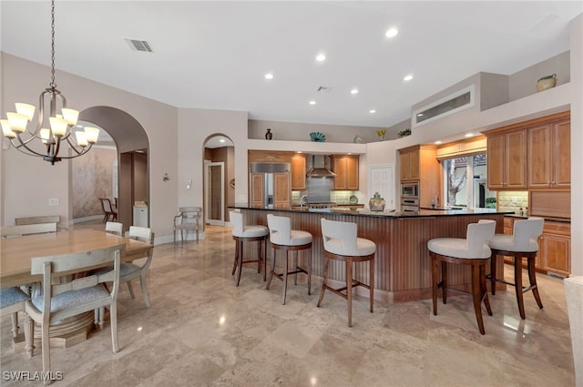 kitchen featuring wall chimney range hood, a kitchen breakfast bar, a notable chandelier, backsplash, and built in appliances