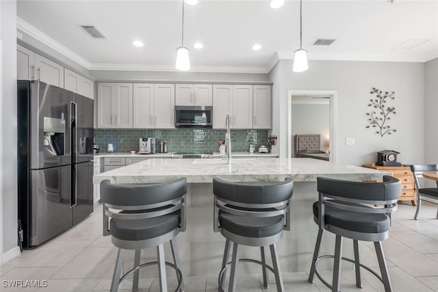 kitchen with a center island with sink, hanging light fixtures, gray cabinets, appliances with stainless steel finishes, and light stone counters