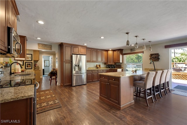 kitchen with appliances with stainless steel finishes, decorative light fixtures, a center island, light stone countertops, and dark wood-type flooring