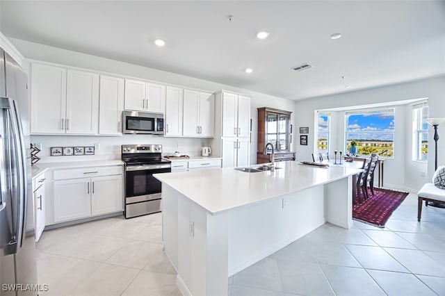 kitchen with stainless steel appliances, sink, light tile patterned floors, white cabinets, and an island with sink