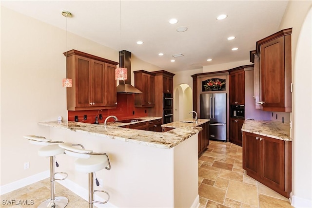 kitchen with kitchen peninsula, appliances with stainless steel finishes, light stone counters, wall chimney exhaust hood, and pendant lighting