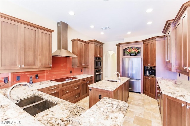 kitchen featuring a center island with sink, sink, wall chimney exhaust hood, appliances with stainless steel finishes, and light stone counters