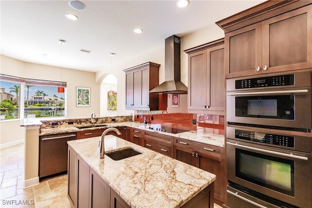 kitchen featuring sink, wall chimney range hood, light stone counters, pendant lighting, and appliances with stainless steel finishes