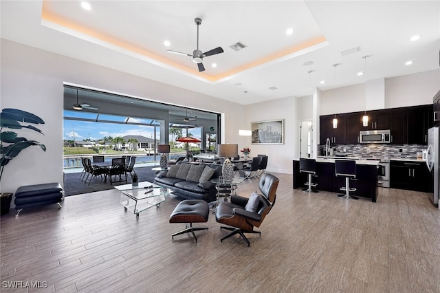 living room with ceiling fan, a tray ceiling, sink, and light hardwood / wood-style flooring