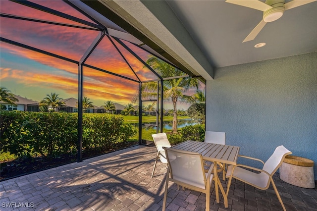 patio terrace at dusk featuring glass enclosure, ceiling fan, and a water view