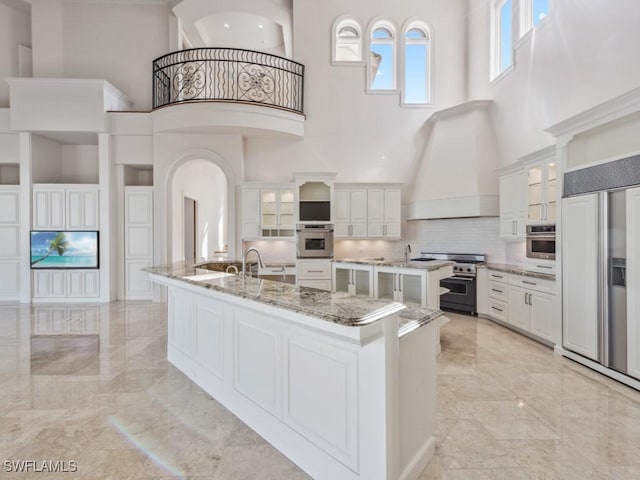 kitchen with high quality appliances, marble finish floor, a high ceiling, custom exhaust hood, and a large island with sink