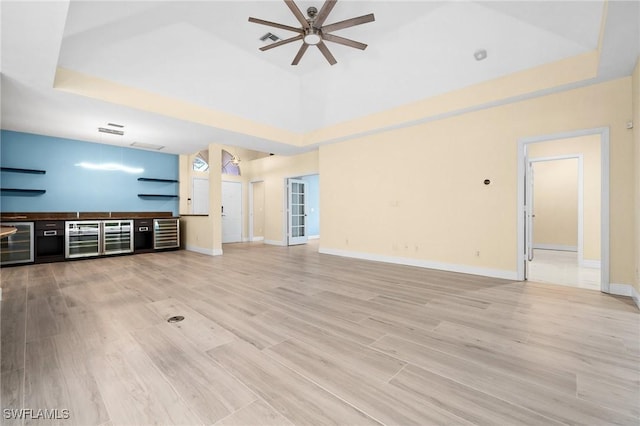 unfurnished living room featuring a raised ceiling, wine cooler, ceiling fan, and light wood-type flooring