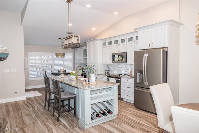 kitchen featuring appliances with stainless steel finishes, decorative light fixtures, white cabinets, light stone counters, and a center island with sink