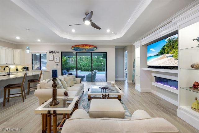 living room featuring a raised ceiling, a glass covered fireplace, crown molding, and light wood finished floors