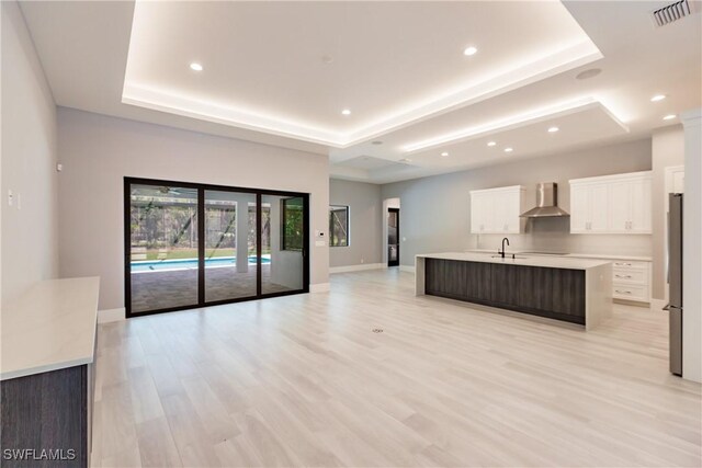 kitchen with white cabinets, wall chimney exhaust hood, a raised ceiling, and a kitchen island with sink