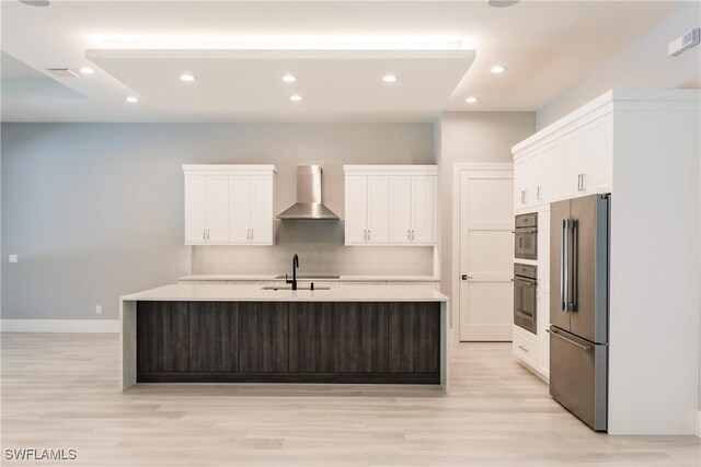 kitchen featuring a center island with sink, white cabinets, wall chimney range hood, and appliances with stainless steel finishes