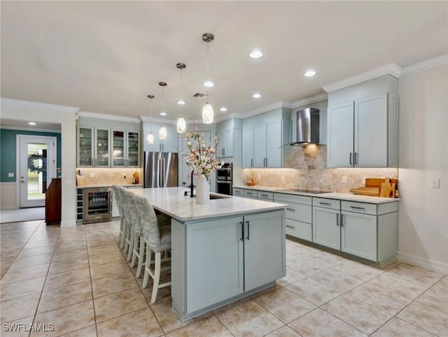 kitchen with a center island with sink, wall chimney range hood, hanging light fixtures, stainless steel appliances, and beverage cooler