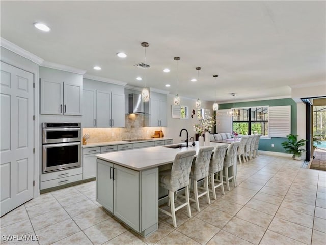 kitchen with sink, double oven, wall chimney exhaust hood, and an island with sink