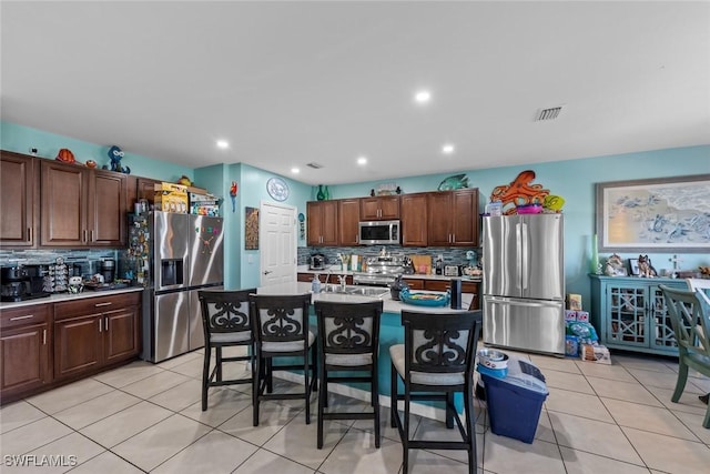 kitchen featuring light tile patterned floors, a breakfast bar area, backsplash, stainless steel appliances, and a center island with sink