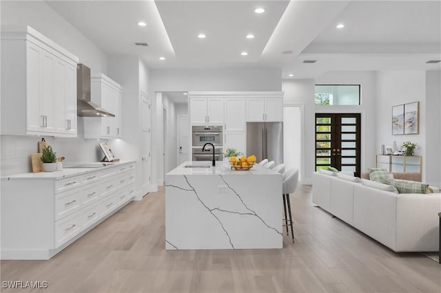 kitchen featuring sink, wall chimney exhaust hood, stainless steel appliances, a large island with sink, and white cabinets