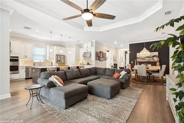 living room with light wood-type flooring, a tray ceiling, ceiling fan, crown molding, and sink