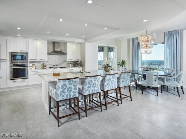 kitchen with a raised ceiling, wall chimney exhaust hood, an island with sink, appliances with stainless steel finishes, and a kitchen bar