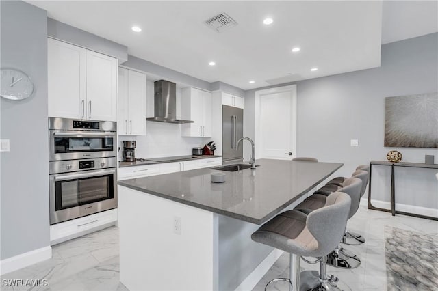 kitchen with white cabinets, wall chimney range hood, sink, an island with sink, and stainless steel double oven